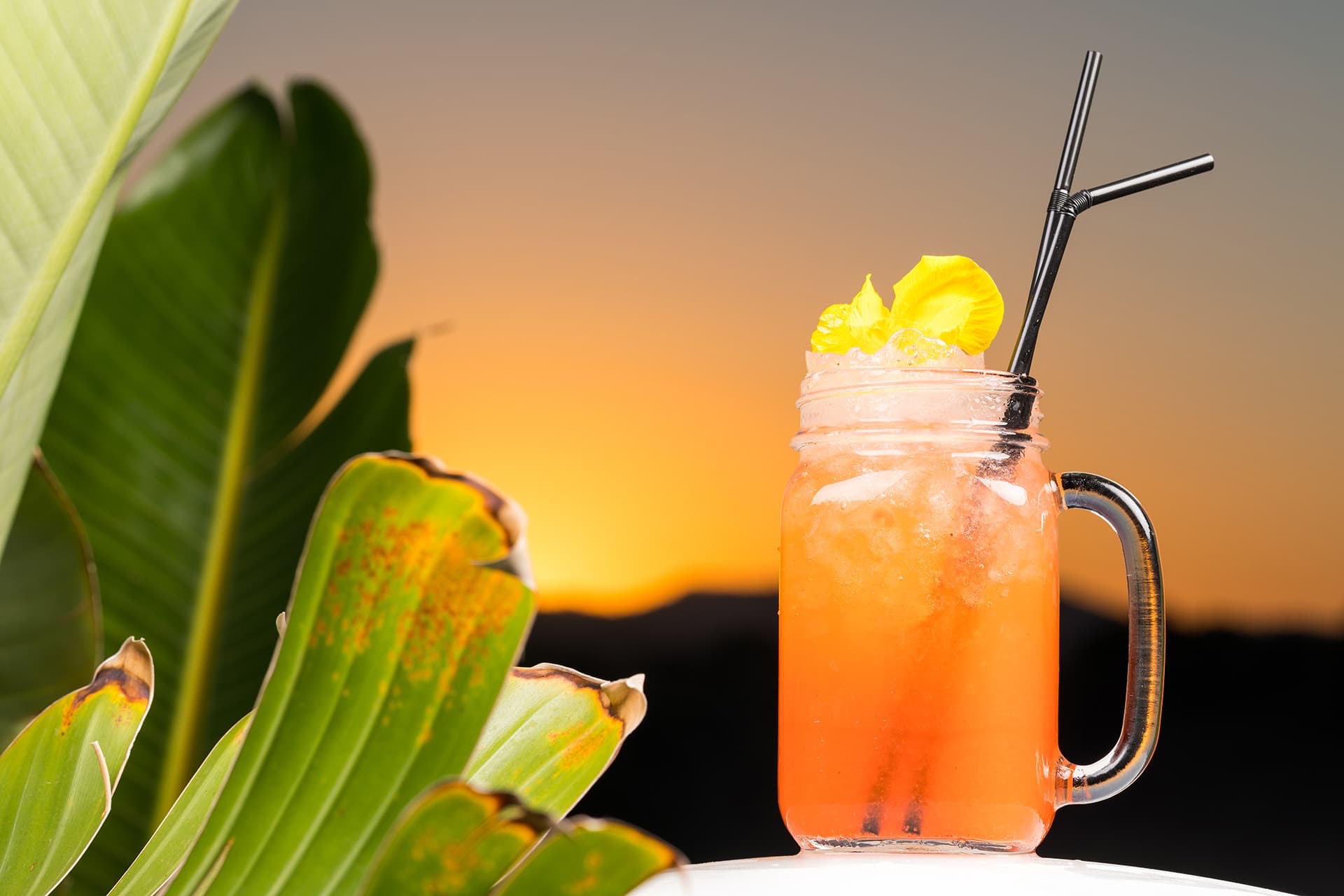 A coral cocktail in a frosted glass beside a banana-leaf plant on the Monsuau rooftop, the Cala d'Or hills a dark line against a gold and charcoal sunset