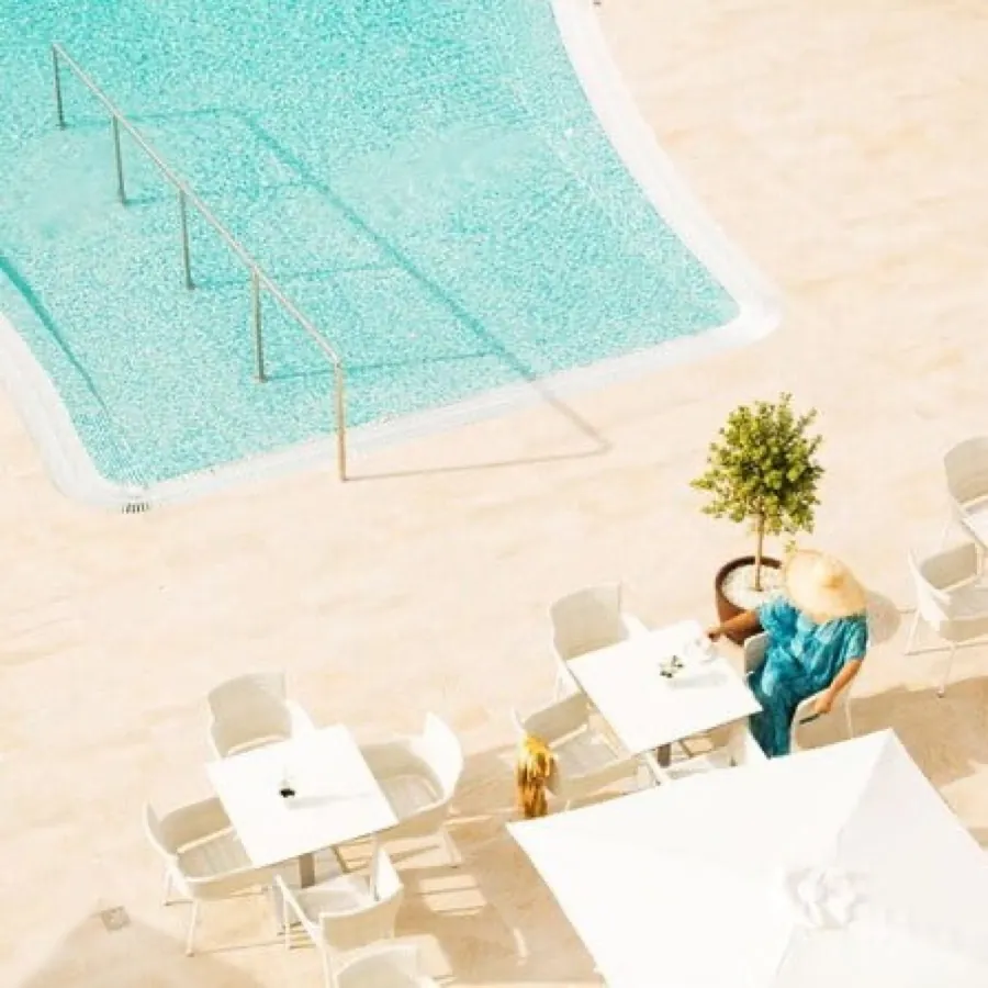 Seen from above: the turquoise Monsuau pool in pale travertine, a woman in a sun hat at a white table by a potted lemon tree, hard noon shadows across the deck