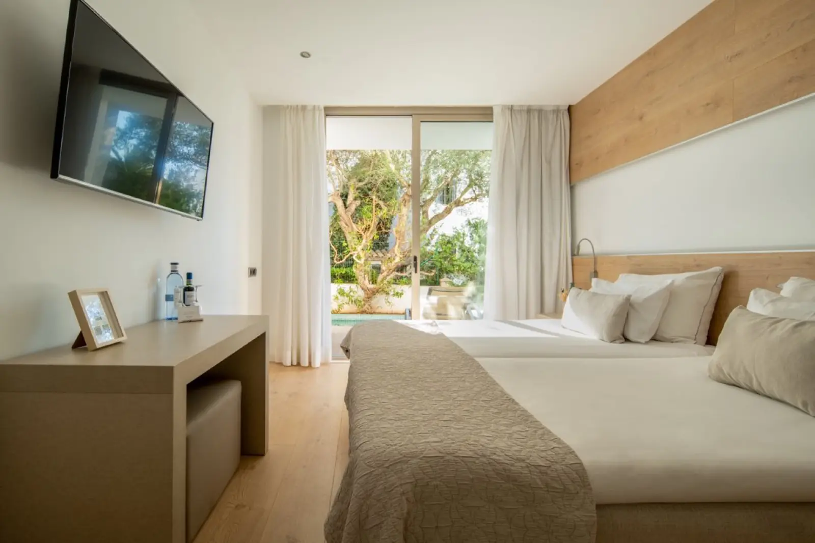 A bright White room with a pale oak headboard, soft green artwork above the bed and a full-height window onto greenery