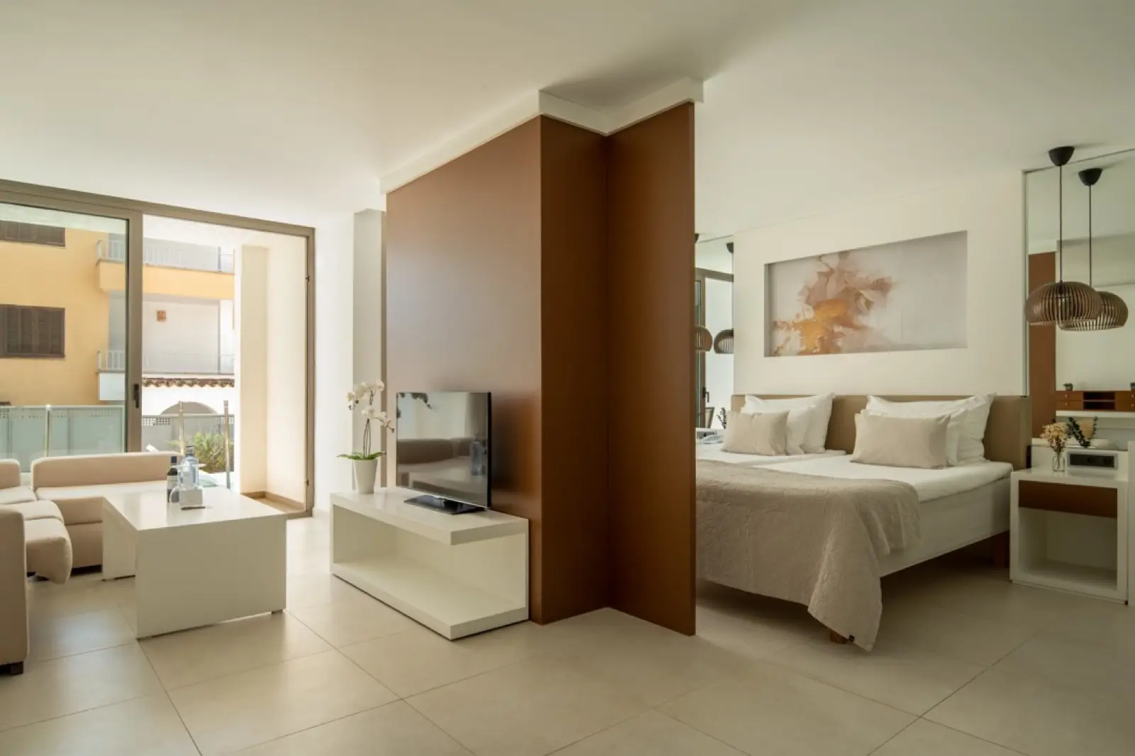 A Junior Suite with a separate sitting area, a tan partition wall, cream sofas and a bed beyond, opening to a sunny balcony